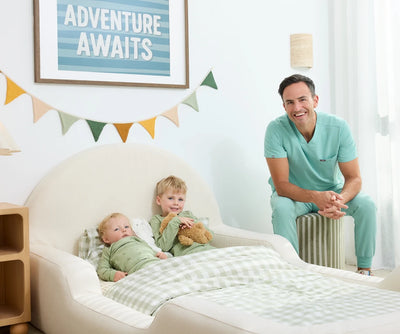 A smiling man in mint green medical scrubs sits on a small stool in a bright nursery, looking toward two babies lying in a cream-colored bassinet. The room has a light wall, a framed 'ADVENTURE AWAITS' sign, a string of colorful flags, and a green-and-white checkered blanket.