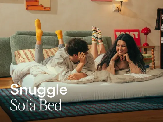 Two people lying on a sofa bed in a living room with 'Snuggle Sofa Bed' text.
