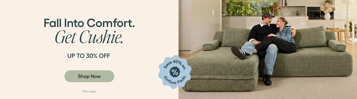A couple relaxes on a large olive green sectional sofa in a modern living room setting, with promotional text on the left reading 'Fall Into Comfort. Get Cushie.