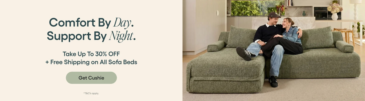 A promotional banner for the Cushion brand showing a man and woman sitting on a large, plush olive green sectional sofa in a living room setting. The left side features text that reads 'Comfort By Day.