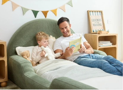 A man and a young child relax in a large green upholstered bed reading a book in a bright, modern bedroom.